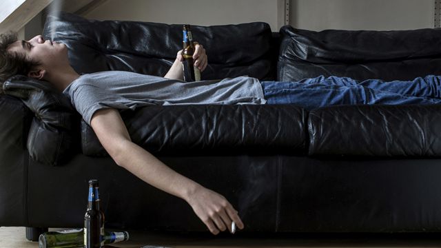 A young man lying on the couch eating beer and smoking