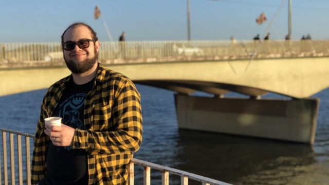 Brazilian cosmologist Pedro Bernardinelli in front of a bridge in the United States drinks coffee.  He wore a plaid shirt (yellow and black) and a red shirt underneath;  He wears sunglasses.