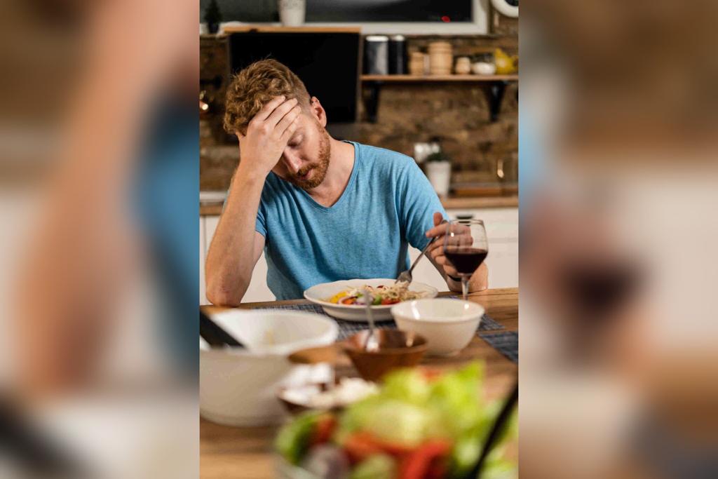 In the color image, the person is placed in the middle.  He is sitting on a chair in front of a table with food.  He has one hand on his head as he looks at a plate of food